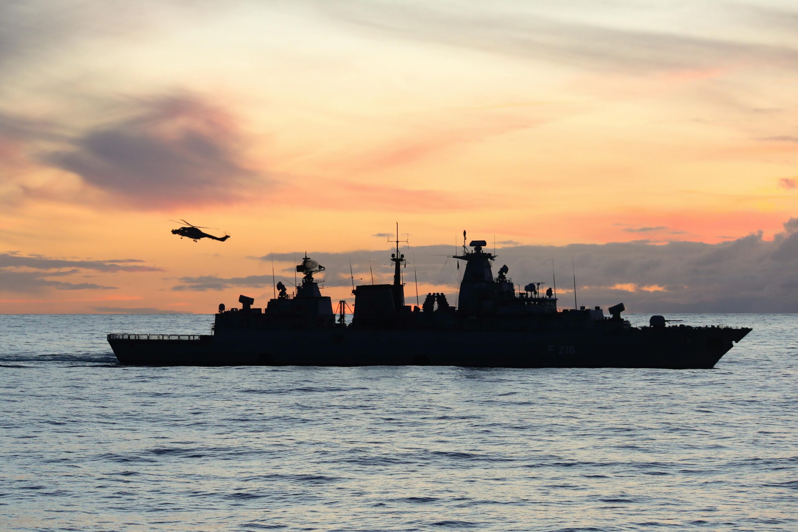 Silhouette of a warship and helicopter at sea during sunset.